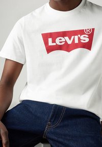Bílé tričko s krátkým rukávem s velkým červeným logem Levi's ve tvaru oblouku. Nosit s tmavě modrými džíny, které zvýrazňují texturu denimu.