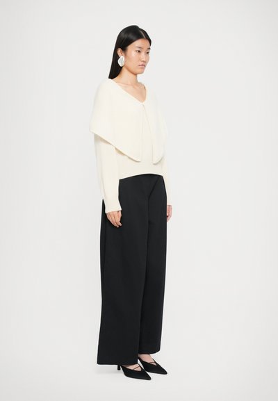 Pull crème avec un col drapé, associé à un pantalon large noir et des escarpins noirs à bout pointu. Texture de tissu lisse.