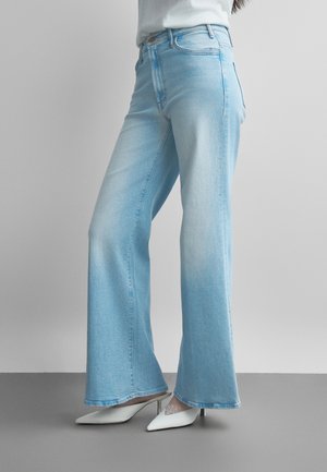 Osoba nosiaca svetlomodré rozšírené rifle a biele špicaté vysoké podpätky, stojaca pred jednoduchým sivým pozadím.