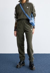 Olivgrön fleece-träningsoverall med vita ränder, utrustad med dragkedja i kragen och elastisk midja. Tillsammans med svarta kraftiga snörstövlar och en blå väska.