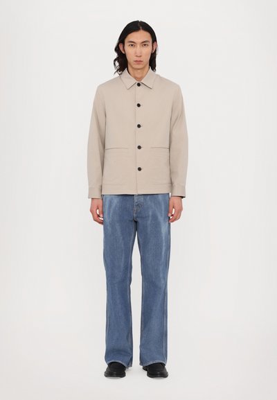 Jachetă din bumbac bej cu guler, nasturi negri și două buzunare frontale, asortată cu blugi din denim deschis albastru cu croială largă și pantofi negri.