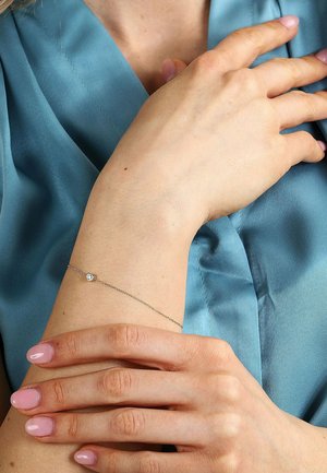Silberne Kettenarmband mit einem kleinen runden Anhänger. Getragen an einem Handgelenk mit heller Hautfarbe, dazu pastellrosa manikürte Nägel und ein Hintergrund aus blauer Satinstoff.