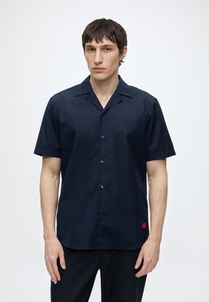 Homme portant une chemise bleu marine à manches courtes avec col, debout devant un fond clair uni.