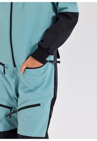 Een teal en zwarte jumpsuit met een rits aan de voorkant, verstelbare manchetten en meerdere ritszakken aan de zijkanten en op de dijen.