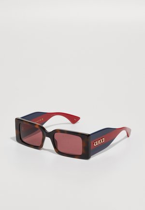 Óculos de sol quadrados com uma armação de tartaruga, hastes vermelhas e azuis, e lentes escuras. Apresenta um logótipo dourado na lateral.