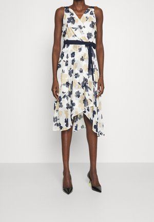 Biała sukienka bez rękawów z granatowo-beżowym kwiatowym wzorem, granatowym paskiem w talii i asymetrycznym dołem, noszona z czarnymi szpilkami o ostrym czubku.