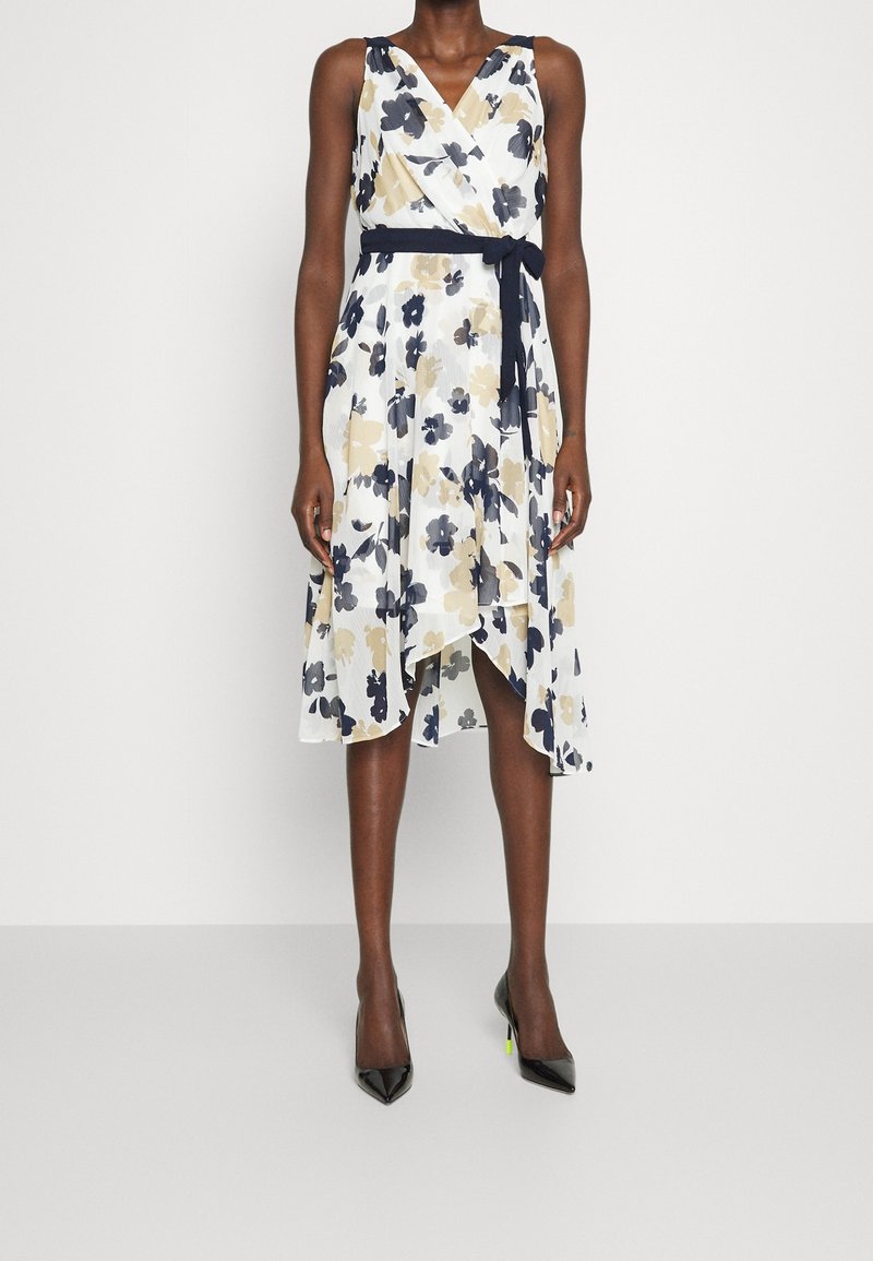 Robe blanche sans manches avec imprimé floral bleu marine et beige, ceinture à la taille bleu marine et ourlet asymétrique portée avec des talons pointus noirs.