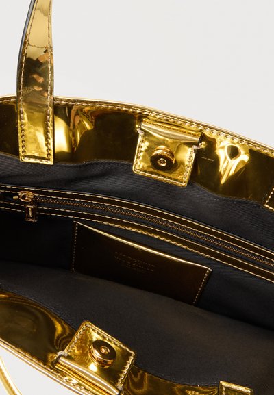 Εσωτερικό μεταλλικής χρυσής τσάντας tote που δείχνει μαύρη υφασμάτινη φόδρα, μία τσέπη με φερμουάρ και μαγνητικούς κλειστούς στα πάνω άκρα.