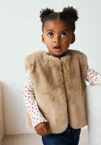 Ung senare med lockigt hår i puffar, bär en beige fuskpälsväst, långärmad tröja med prickar och mörka jeans, står med en hand på en yta.