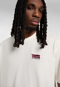 Sweatshirt blanco de cuello redondo hecho de algodón suave, con un pequeño logo bordado en burdeos y azul que dice "Dickies Workwear" en el pecho.