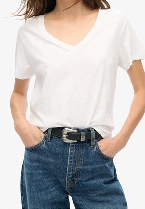 Femme portant un t-shirt blanc à col en V rentré dans un jean bleu avec une ceinture noire ornée d'une boucle décorative argentée. Mains dans les poches.