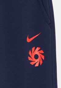 Tumši zilie sporta bikses ar sarkano Nike logotipu un grafisku spirālveida dizainu, izgatavotas no mīksta kokvilnas maisījuma auduma, ar sānu kabatām.