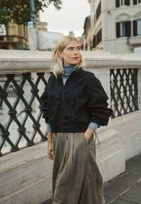 Mladá žena s blond vlasmi v čiernej bunde a sivej sukni stojí pri kamennom zábradlí v mestskom prostredí, pozerá sa doprava.