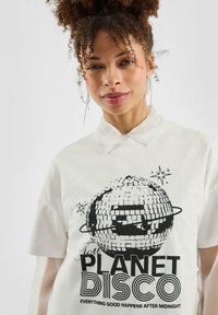 Witte oversized t-shirt met een zwart grafisch ontwerp van een discobal en de tekst "Your PLANET DISCO" met sterren. Korte mouwen en kraagdetail.