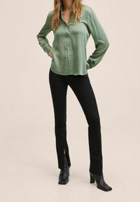 Camicia verde in satin con parte anteriore con bottoni, maniche lunghe e orlo curvo; abbinata a pantaloni neri slim-fit con fessure laterali.