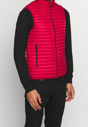 Gilet rembourré rouge avec un col montant, design matelassé horizontal, deux poches latérales zippées et une fermeture éclair à l'avant. Porté par-dessus une chemise noire à manches longues.