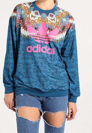 Kobieta ubrana w niebieską, wzorzystą bluzę Adidas z różowym logo i podartymi niebieskimi dżinsowymi szortami, stojąca na jednolitym tle.