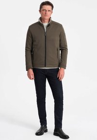 Giacca aderente verde oliva con colletto alto, apertura con zip, due tasche con zip, tessuto testurizzato, indossata sopra un top grigio chiaro e jeans scuri.