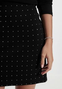 Jupe noire au toucher doux ornée de cristaux en argent disposés en motif, mise en valeur par une main portant un bracelet en argent.