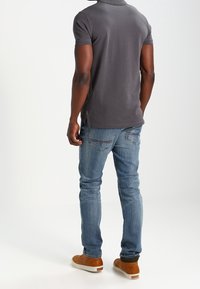 Man i mörkgrå pikétröja, blå jeans med söm på bakfickan, och bruna vardagsskor som står med ryggen vänd.