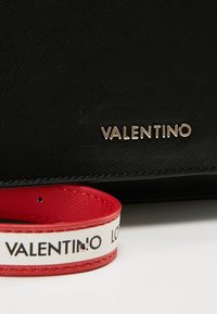 Sac Valentino noir texturé avec logo doré et une sangle rouge et blanche arborant le nom Valentino en lettres noires.