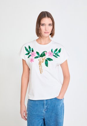 Mujer vistiendo una camiseta blanca con un diseño de loro de lentejuelas, hojas verdes y flores rosas, combinada con pantalones vaqueros azules, de pie sobre un fondo blanco.
