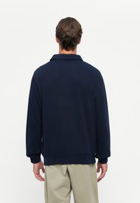 Mann steht mit dem Rücken zur Kamera, trägt einen dunkelblauen Langarm-Pullover mit Kragen und beige Hosen vor einem schlichten weißen Hintergrund.
