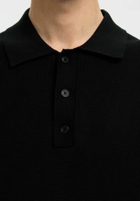 Gros plan d'un polo noir texturé avec un col et trois boutons noirs, porté par une personne.