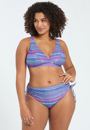 Kvinde med krøllet hår iført en blå og lyserød mønstret bikini, som kigger let til siden med den ene hånd på hoften.