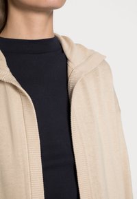 Beżowy dzianinowy kardigan z kapturem, z prążkowanym wykończeniem, noszony na ciemnej koszuli, prezentujący gładkie tekstury i luźny fason.