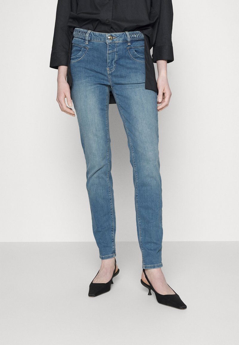 Personne portant un jean bleu slim associé à des chaussures slingback à petit talon noir et une chemise noire à manches longues.