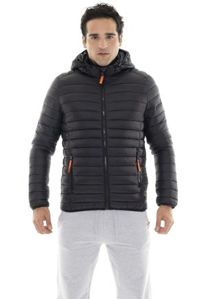 Un homme porte une veste noire matelassée à capuche avec des détails orange au niveau des fermetures éclair et un pantalon de jogging gris clair, sur fond blanc.