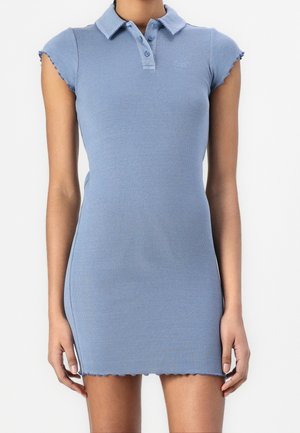 Femme portant une robe polo moulante bleu clair à manches courtes côtelées avec un col et une patte de boutonnage, debout devant un fond blanc uni.