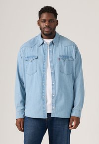 Hele sinine teksasärk pikkade varrukatega, nööbiga ees, kahe rinnataskuga ja terava kraega. Kandes valge t-särgi kohal.