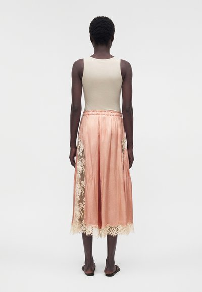 Άτομο που φοράει ένα αμάνικο μπεζ τοπ και μια ροζ σατέν φούστα με δαντελένιες λεπτομέρειες, που στέκεται ξυπόλητο σε απλό λευκό φόντο.