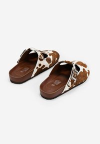 Zuecos de estampado de vaca marrón y blanco con una correa ajustable, un upper de textura suave y una suela de goma marrón.