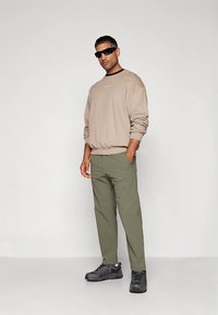 Bézs pulóver fekete gallérral, olíva zöld nadrág és fekete sportcipő. A modell napszemüveget visel, semleges háttér előtt áll.