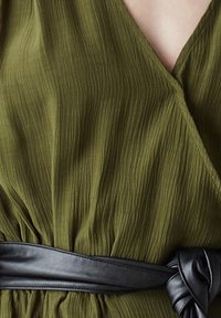 Robe portefeuille texturée vert olive avec un décolleté en V profond, cintrée à la taille par une ceinture en cuir noir nouée.