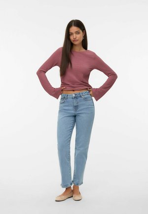 Eine Frau mit langen, dunklen Haaren trägt ein strukturiertes, mauvefarbenes Langarmoberteil, hellblaue Jeans und beigefarbene Ballerinas. Sie steht mit den Händen in die Hüften gestützt vor einem weißen Hintergrund.