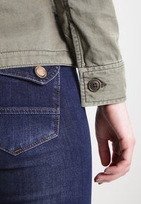 Oliven grøn jakke med en struktureret finish, der har en manchet med knapper. Mørkeblå jeans med synlige sømme og en laplomme.