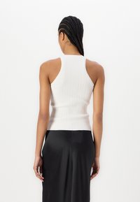 Mujer con cabello trenzado que lleva una blusa sin mangas de canalé blanca y una falda negra, vista de espaldas contra un fondo blanco liso.