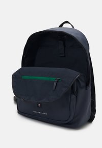 Zaino blu navy con pattina anteriore, tasca con cerniera verde, dettaglio del logo e spallacci imbottiti. Realizzato in materiale durevole, presenta un interno spazioso.