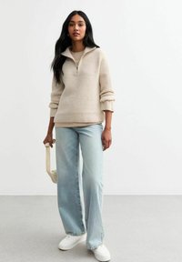 Pull zippe beige avec des côtes texturées, associé à un jean large bleu clair et des baskets blanches. Tient un petit sac de couleur claire.