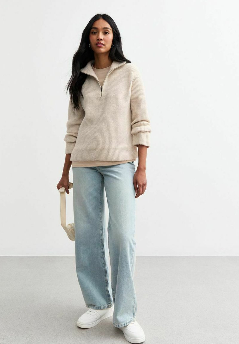Pull zippe beige avec des côtes texturées, associé à un jean large bleu clair et des baskets blanches. Tient un petit sac de couleur claire.