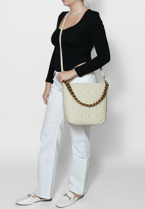 Beige gewatteerde handtas met een gouden kettingband, met een gladde textuur en geometrisch patroon. Draagt met een zwarte top en witte broek.