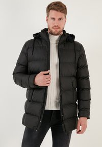 Melna pūkaina jaka ar rāvējslēdzēju, quilted dizainu un kapuci. Materiāls ir gluds ar minimālu šuvju detalizāciju.