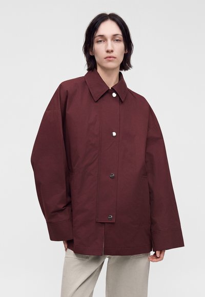 Personne aux cheveux courts et foncés portant une veste boutonnée rouge foncé oversize et un pantalon beige clair, debout devant un fond uni.