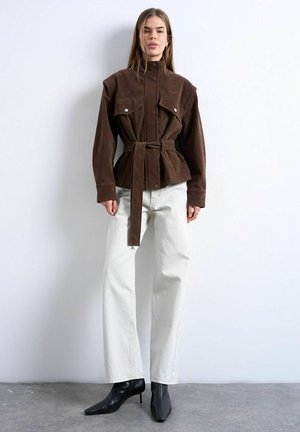 Mujer de pie contra una pared blanca, vistiendo una chaqueta marrón con cinturón, pantalones blancos de pierna ancha y botas de tacón puntiagudo negras hasta el tobillo.