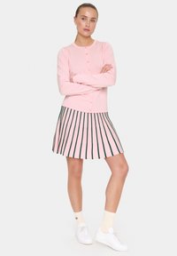 Cardigan rose à manches longues avec fermeture à boutons, associé à une jupe plissée avec des rayures roses et vertes. Baskets blanches et chaussettes crème.