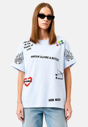 Jonge vrouw die een lichtblauw oversized T-shirt draagt met zwarte tekst en grafische prints, waaronder dobbelstenen, hart met "Karma," en kleine kleurrijke pictogrammen.